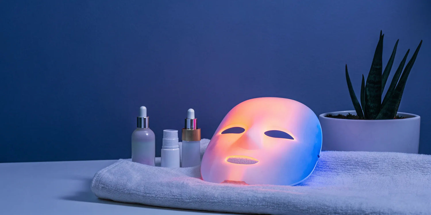 A silicone LED mask glows with red light next to various skincare products.